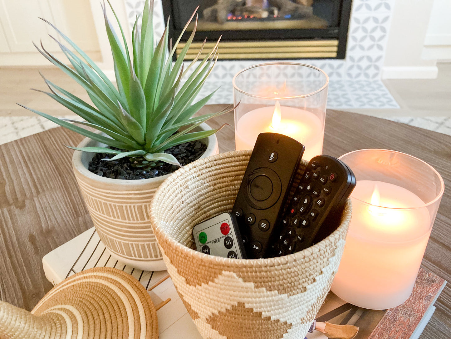 woven basket with white zig zag pattern holding remote controls on coffee table.  There is also a faux succulent plant and 2 candles. All items are on top of a coffee table book for display