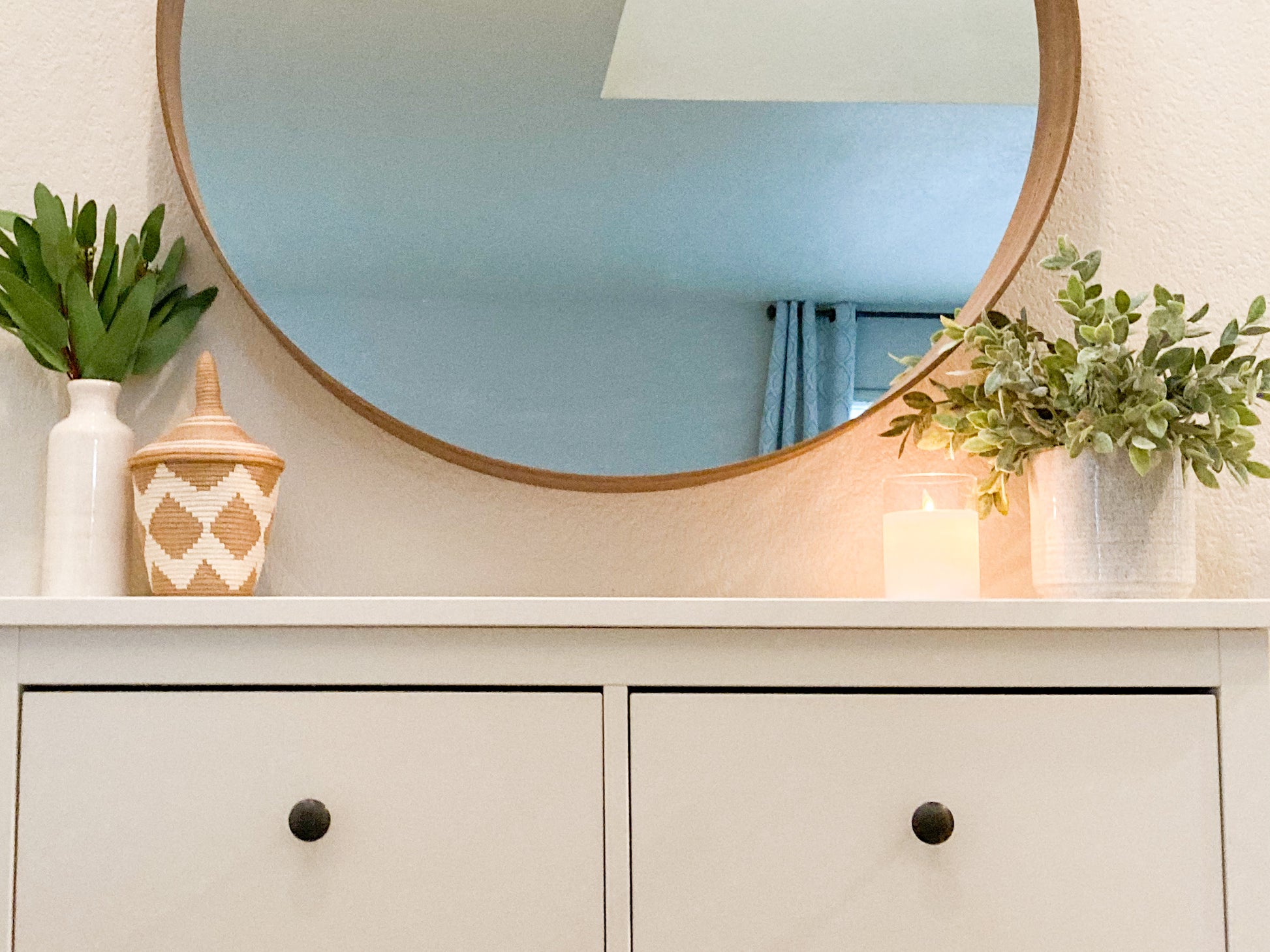 entryway table with faux leaf arrangement in a white bud vase, a small woven basket with a pointed lid, a candle and another faux green plant. A large round mirror is above the entryway table.