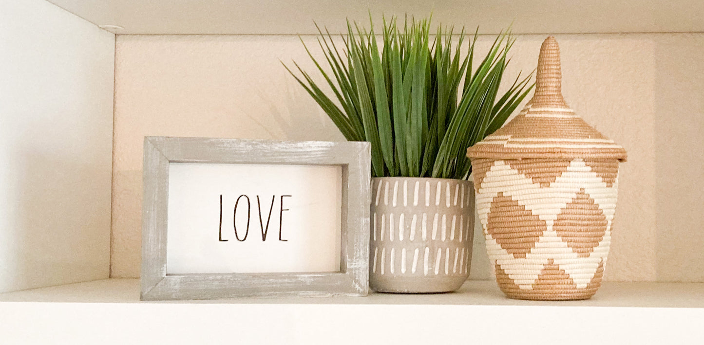 Love sign with gray frame, faux green grass plant, hand woven basket with white zig zag pattern on the shelf